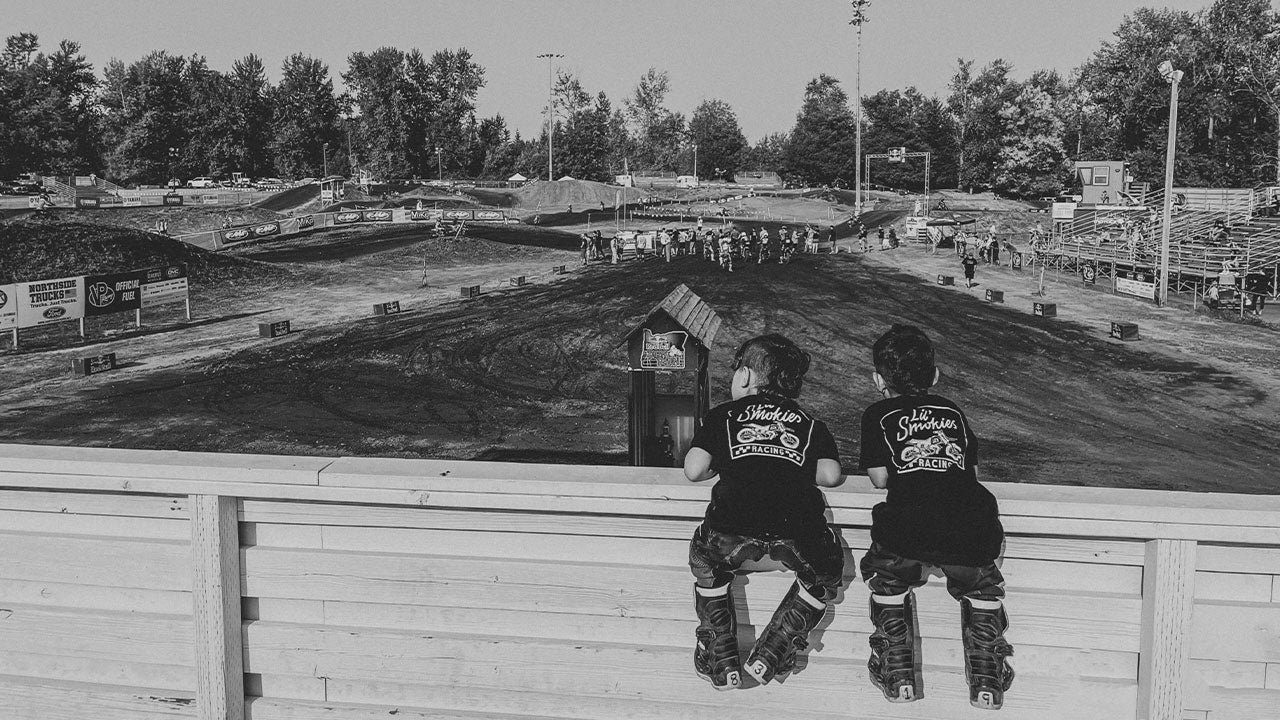 Image of two youth motocross racers watching dirt bikes race at thursday night motocross. The kids are leaning over a fence wearing lil' smokies racing apparel and moto gear. 