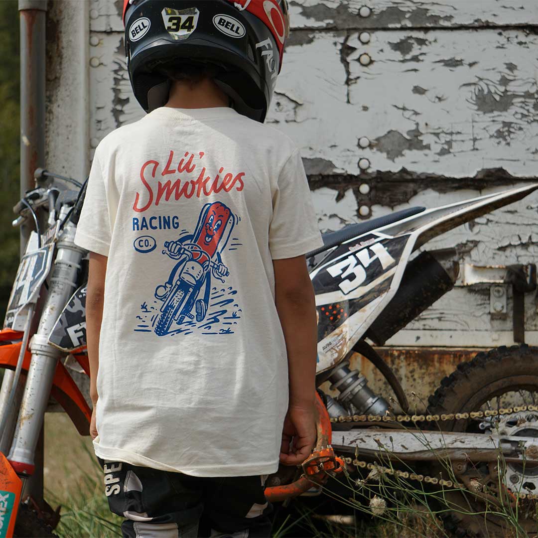 On body image of a youth wearing the Lil Smokies Racing Burt dogs tee standing with a dirt bike helmet on next to a orange dirtbike. Image shows the detail of the design and the fit of the tee. Model is wearing a Youth Medium