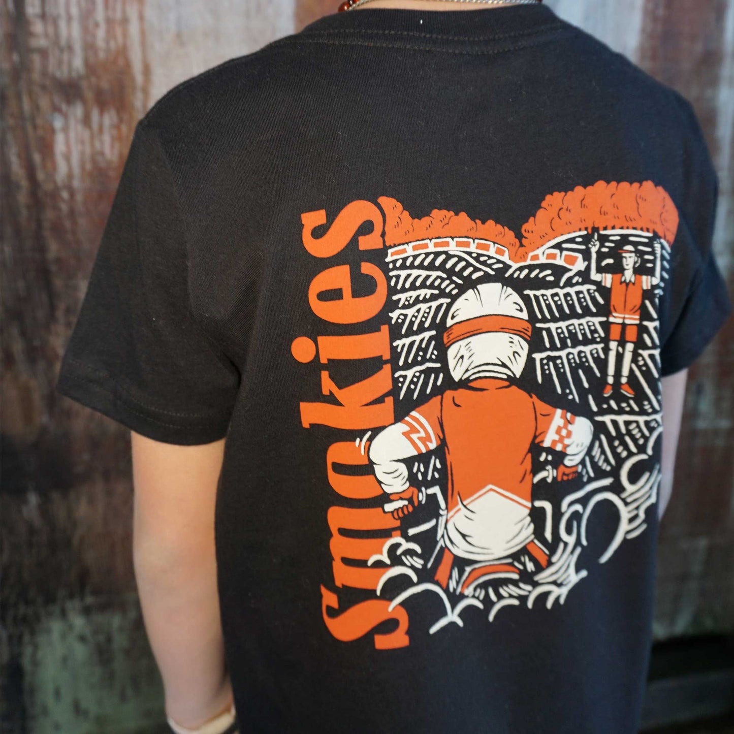 On body image of the back graphic of the Smokies black tshirt in youth sizing. Full graphic on the back with the word Smokies written vertical on the left in red with a dirt bike rider elbows up at the starting gate waiting for the starter to come back and drop the gate.
