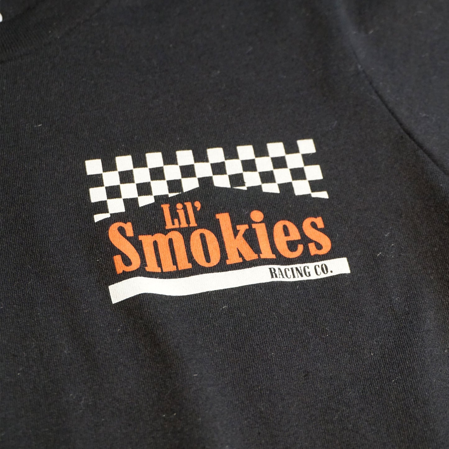 Close up of the front image of the youth Smokies tshirt showing the left front chest with a checkered black and white flag with the words Lil Smokies in red ink.