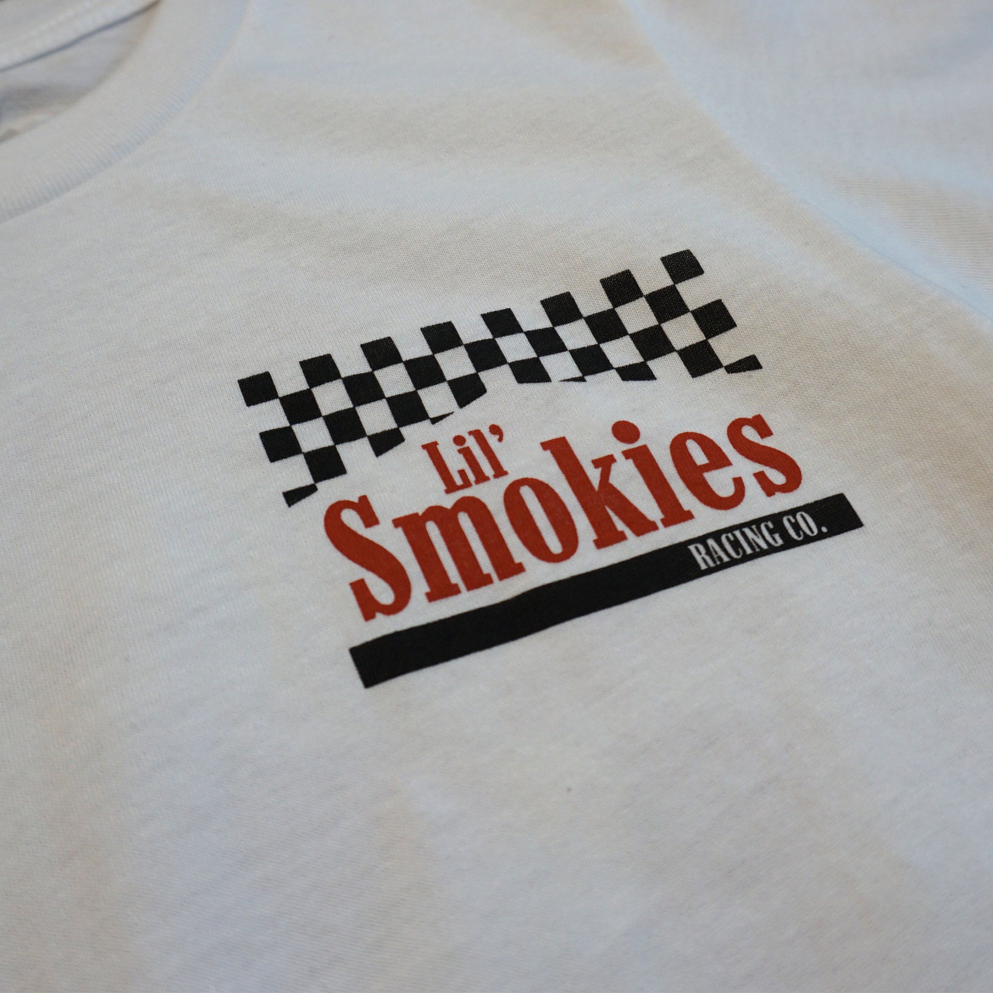 Close up of the front image of the youth Smokies tshirt showing the left front chest with a checkered black and white flag with the words Lil Smokies in red ink.