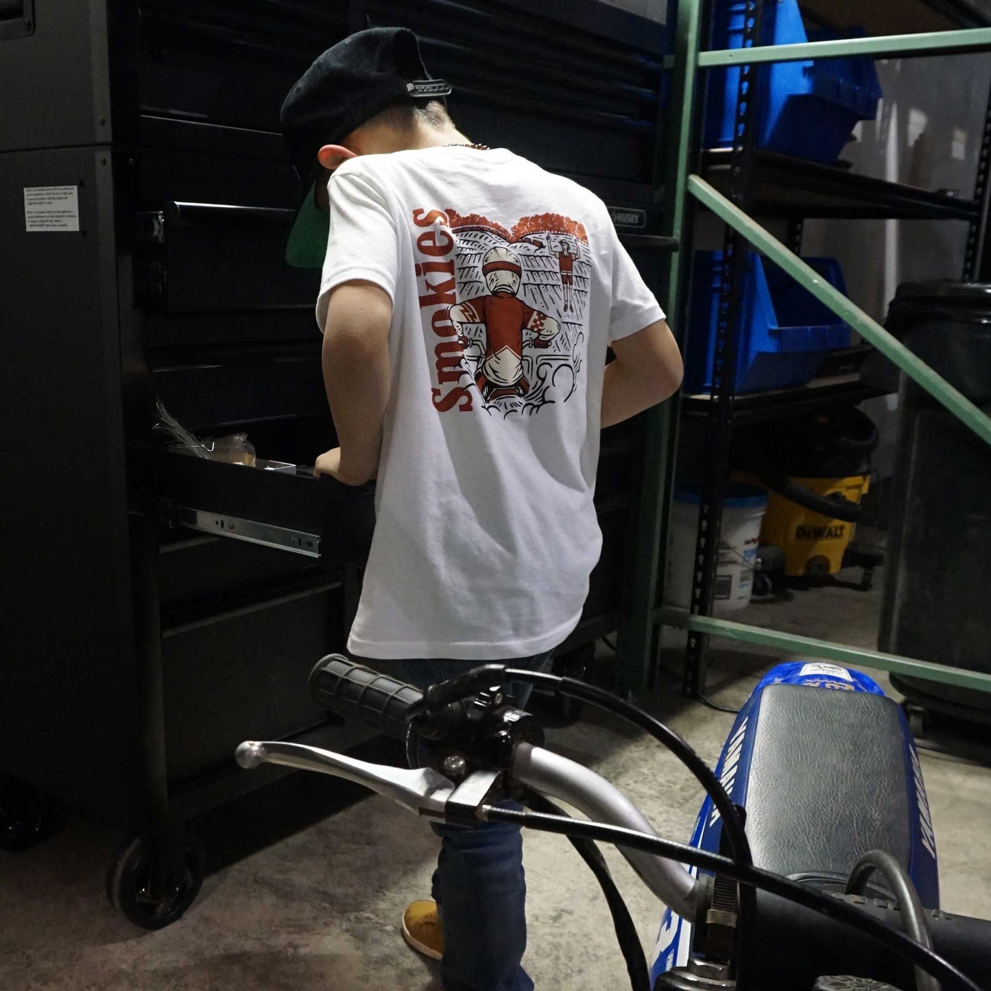 Capture the essence of motocross adventure with our dynamic t-shirt, featuring a fearless racer amidst the action. Perfect for young riders and enthusiasts, this tee embodies the thrill of the track. Inspire the next generation of motocross champions with our premium quality t-shirt. Crafted for comfort and durability, it's the ideal choice for kids who love to ride. Shop now and ignite the passion for motocross in your little one's wardrobe. Rider working on his Yamaha PW 50 in the garage shop.