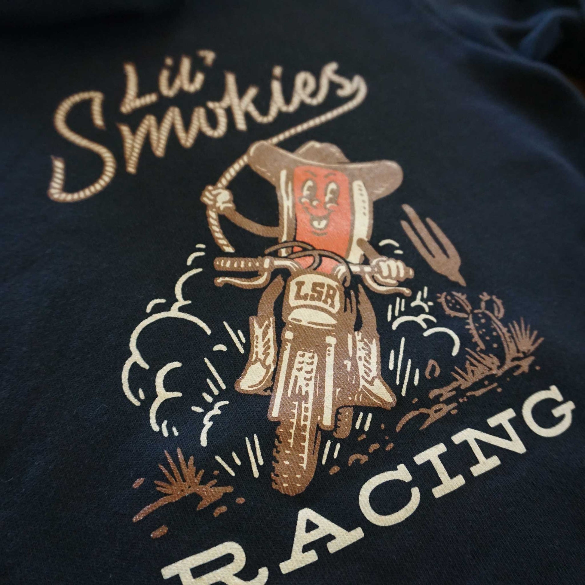 Close up image of the Cowboy hotdog riding a dirtbike in the desert. Image in brown, tan and burnt orange on a black hoodie.