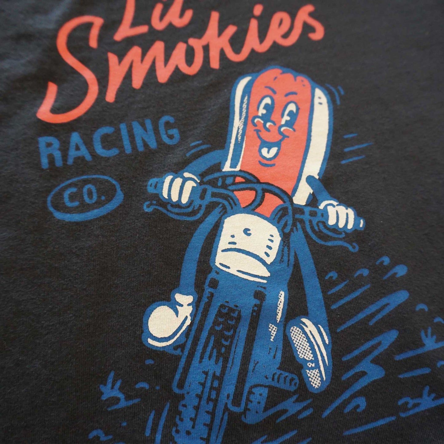 Close up image of the back of the tee. Youth black short sleeve tshirt with Lil' Smokies racing font logo. A hotdog in a bun riding a motorcycle with a big smile on its face and dirt flying up from the tires. Playful cartoon image